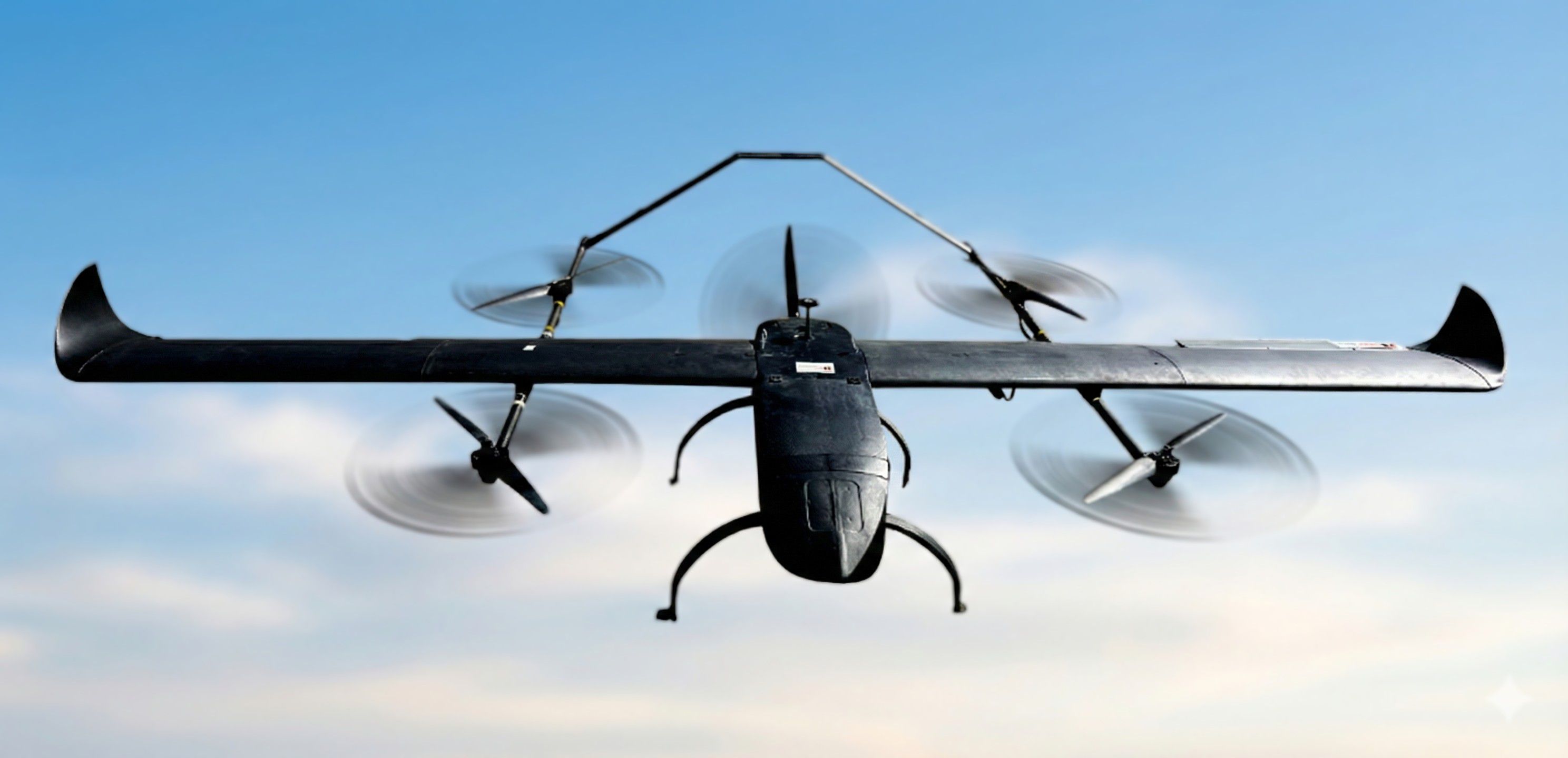 Front view of a black fixed-wing VTOL drone featuring four vertical lift propellers and a rear pusher propeller, set against a smooth, soft-focused sky background.